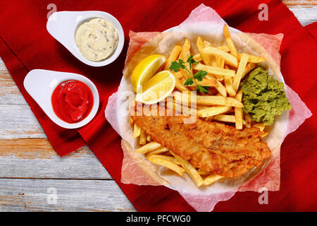 Poisson et frites croustillantes - Cabillaud frit, frites, rondelles de citron, de sauce tartare et purée de petits pois sur la plaque sur le papier sur la vieille table en bois , recette authentique Banque D'Images