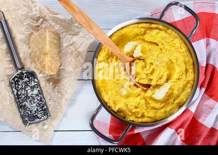 La polenta chaude délicieux porridge avec du beurre et du fromage râpé dans la poêle avec une cuillère de bois, une râpe avec morceau de fromage parmesan sur papier sur le tableau blanc, Banque D'Images