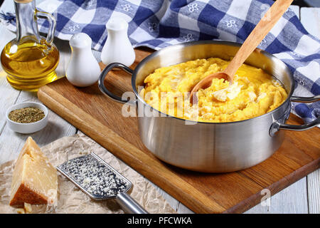 Délicieux porridge chaud de base de la polenta avec du beurre dans la poêle avec une cuillère de bois sur une planche à découper, de parmesan, une râpe. Bouteille d'huile d'olive, de sel et de pep Banque D'Images