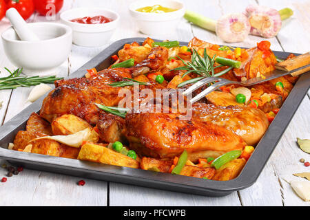 La peau du poulet grillé croustillant d'or sur la plaque de cuisson avec les pommes de terre, ail,. les pois, haricots verts, carottes.et fourchette à découper en bois blanc sur les ingrédients. Banque D'Images