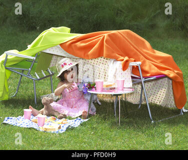 Tente de fortune faits de chaises et couvertures, fille assise sous tente, de porter un chapeau et d'alimentation de nounours. Banque D'Images