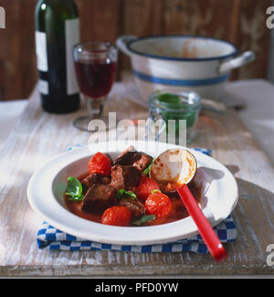 Agneau provencale, morceaux de viande, les tomates cerise, les feuilles de basilic dans la sauce tomate, servi en soupe, plat blanc louche embedded, verre, bouteille de vin et casserole en arrière-plan Banque D'Images