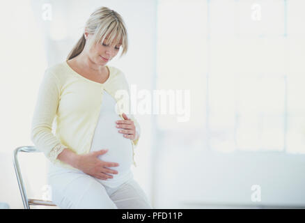 Femme blonde assise à la fin de l'étape de la grossesse à la recherche vers le bas et de caresser son ventre, vue de face. Banque D'Images