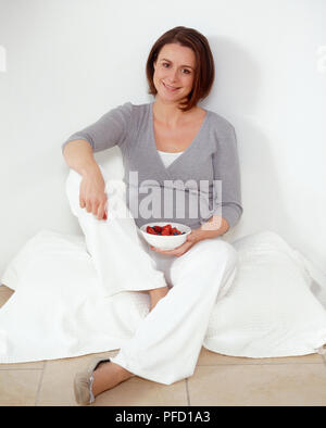 Pregnant woman sitting on coussin sur le sol, tenant bol de fraises, smiling Banque D'Images