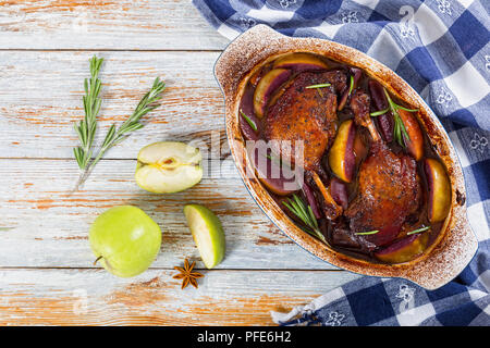 Cuisses de canard rôti cuit au four dans du vin rouge avec le romarin et le zeste d'orange et les tranches de pommes dans un plat à gratin, les étoiles d'anis et de pomme verte sur peelin blanc Banque D'Images