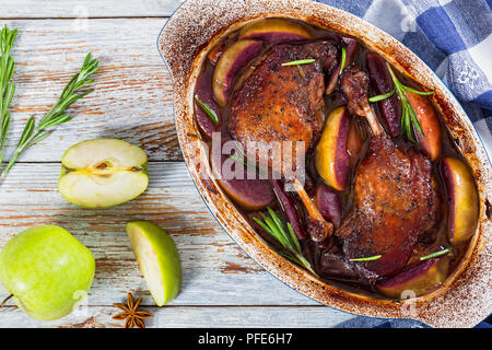 Cuisses de canard rôti cuit au four dans du vin rouge avec le romarin et le zeste d'orange et les tranches de pommes dans un plat à gratin, les étoiles d'anis et de pomme verte sur peelin blanc Banque D'Images