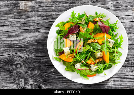 Délicieux sain salade de kaki avec le mélange d'Épinards, roquette et feuilles de laitue avec du fromage bleu et noix sur la plaque blanche sur la vieille planche de bois foncé Banque D'Images