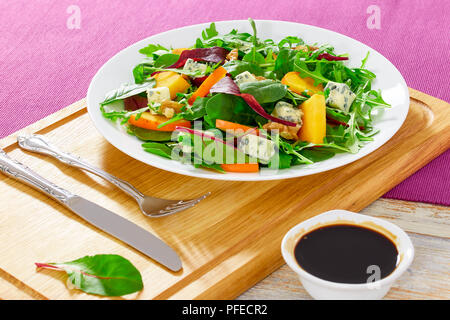 Kaki, fromage bleu, roquette, jeunes épinards, laitue et noix délicieuse salade sur assiette blanche sur planche à découper en bois avec couteau, fourchette, et b Banque D'Images