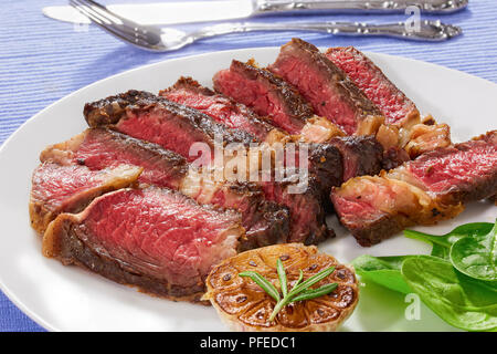 Steak juteux à point boeuf aux épices coupées en tranches sur plaque blanche avec l'ail frit et salade d'épinards frais vert. fourchette et couteau sur arrière-plan, c Banque D'Images