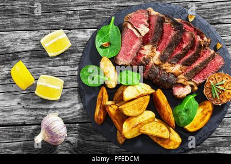 Biftecks juteux steak de boeuf saignante avec épices coupées en tranches sur la plaque en ardoise avec de l'ail frit et frites et salade d'épinards. Les quartiers de pommes de terre, citron et Banque D'Images