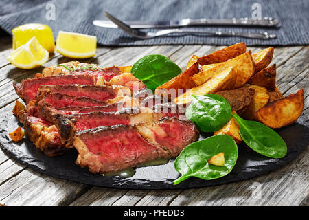 Moyen-grillé Steak faux-marbre rare coupé en tranches sur la plaque en ardoise avec de l'ail frit, salade d'épinards et les quartiers de pommes de terre frites. dark style, voir à partir de Banque D'Images