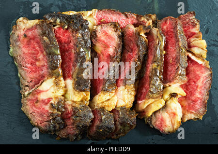 Rosée bifteck de faux-filet grillés aux épices coupées en tranches sur la plaque en ardoise noire répandu avec du beurre l'ail sauce à la viande, close-up Banque D'Images