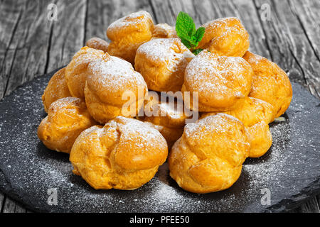 Profiteroles délicieux rempli de crème sur la plaque d'ardoise, saupoudrage de sucre glace et décoré de menthe, sur la vieille table en bois foncé, close-up Banque D'Images