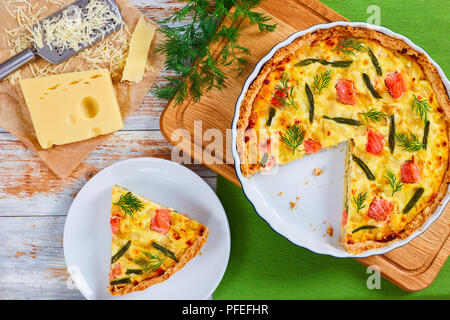 Quiche au saumon, français avec haricots verts et fromage, crème et oignon coupé en tranches dans le plat de cuisson sur la planche à hacher. tranche de la tarte sur une assiette, une râpe et p Banque D'Images