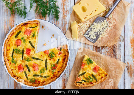 Quiche au saumon, français avec haricots verts et fromage, crème et oignon coupé en tranches dans le plat de cuisson sur la vieille table en bois. tranche de la tarte sur une assiette, une râpe et Banque D'Images