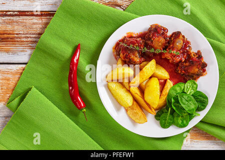 Rabo de Toro ou de boeuf mijotée dans sauce tomate au vin rouge sur plaque blanche servi avec salade et frites de quartiers de pommes de terre, sur la vieille table en bois blanc, au Banque D'Images