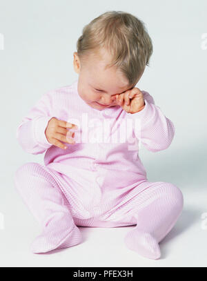 Petite fille en rose tenue assise sur le sol et se frottant les yeux, vue avant Banque D'Images