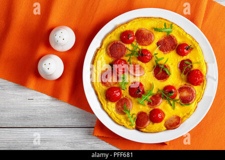 Délicieux omelette avec saucisses coupées en tranches, les tomates cerises entières et roquette fraîche sur plateau blanc sur tapis de table avec salières et poivrières, vi Banque D'Images