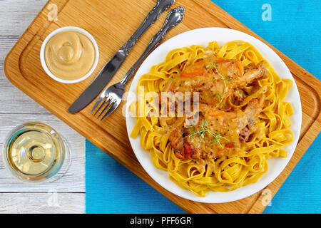 Délicieux lapin braisé ou lapin a la moutarde servi avec des pâtes sur la plaque blanche sur planche de bois avec un verre de vin blanc, recette authentique, vue arrière Banque D'Images