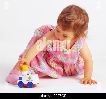 Petite fille à genoux sur le sol, placer de petits poupée en plastique sur le dessus du jouet en plastique blanc ambulance. Banque D'Images