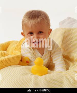 Bébé garçon aux cheveux blonds, sous une couverture, allongée sur le lit, l'estomac lui-même jusqu'à soutenir ses bras, à côté de deux canards en caoutchouc. Banque D'Images