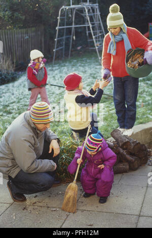 Dans la famille jardin, homme à genoux à côté de garçon enfant en balayant avec balai, woman handing stick à la fille du sac de feuilles, girl raking en arrière-plan Banque D'Images