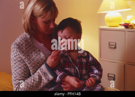 Femme assise avec tout-petit garçon sur ses genoux, holding digital thermomètre dans la bouche du garçon. Banque D'Images