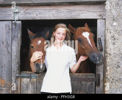 Smiling girl de caresser la tête des deux chevaux (Equus caballus) peeking plus stable porte de la cabine, looking at camera, front view Banque D'Images