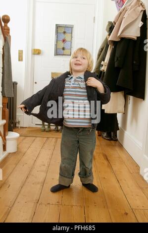 Boy wearing coat, jeans et t-shirt à rayures, standing in hallway Banque D'Images