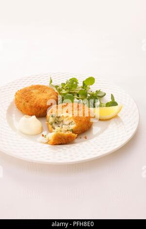 L'aiglefin et herb fishcakes enrobées de chapelure, servi avec le cresson, tranche de citron et la mayonnaise sur une assiette, close-up Banque D'Images