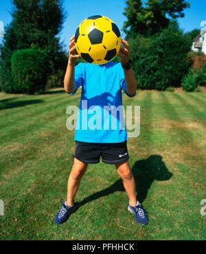 Boy standing in a garden holding un football en plastique en face de son visage Banque D'Images