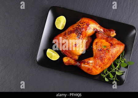 Les cuisses de poulet rôti délicieux croustillant à la peau brun doré sur la plaque noire avec les tranches de citron et feuilles de thym vert frais sur le plateau en ardoise, vue Banque D'Images