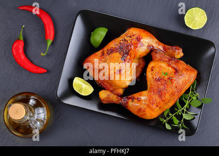 Cuisses de poulet rôti délicieux croustillant à la peau brun doré avec des tranches de citron et feuilles de thym frais vert sur la plaque noire et d'huile d'olive sur withchili Banque D'Images