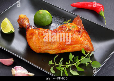 Poulet rôti savoureux quart cuisse avec peau brun doré croustillant avec les tranches de citron et feuilles de thym frais vert sur la plaque noire sur le plateau en ardoise, vue à partir de Banque D'Images