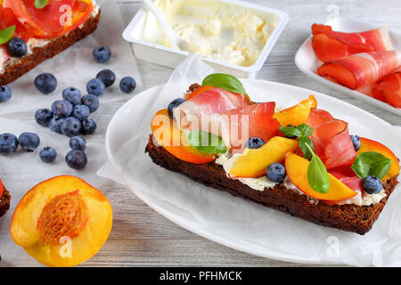 Close-up de délicieux sandwiches ouverts colorés avec du fromage à la crème, de bleuets, de pêche, de tranches de prosciutto et de feuilles de basilic sur le pain de seigle avec des ingrédients Banque D'Images