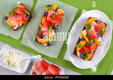 De délicieux sandwiches ouverts colorés avec du fromage à la crème, de bleuets, de pêche, de tranches de prosciutto et de feuilles de basilic sur le pain de seigle avec des ingrédients sur des t Banque D'Images