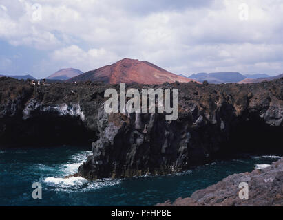 Espagne, Canaries, Lanzarote, El Golfo, Los Hervideros, vagues se briser contre la base de hautes falaises entouré de paysage volcanique au-dessus Banque D'Images