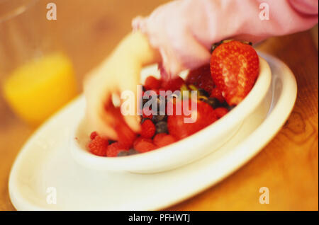 La main de l'enfant hors de framboises un bol de fruits, un verre de jus d'orange en arrière-plan, Close up. Banque D'Images