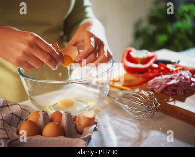 Des gouttes d'œuf cru être shell 12.2005 sur bol en verre, boîte à œufs et fouetter par les côtés de la cuvette, légumes hachés sur planche de bois en arrière-plan. Banque D'Images