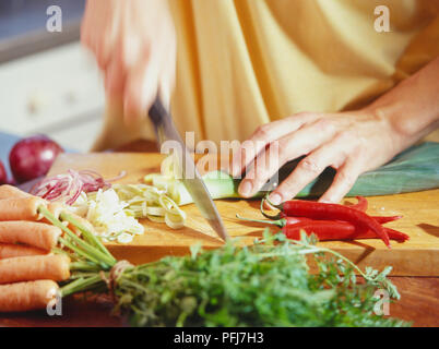 Le poireau étant coupé en tranches sur une planche à découper en bois, à côté de tranches d'oignons rouges, des piments entiers et tas de carottes. Banque D'Images