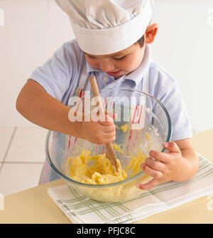 Boy in Chef's Hat battre ensemble le beurre, les oeufs et le sucre dans un bol à mélanger, à l'aide d'une cuillère en bois Banque D'Images