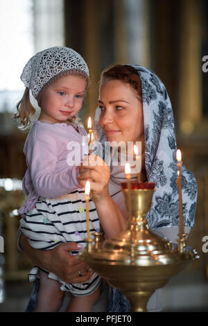 Belle femme russe dans un foulard et aux cheveux rouges tenant une petite fille et s'allume une bougie devant une icône dans l'Eglise orthodoxe russe. Banque D'Images