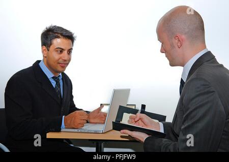 Homme assis avec un ordinateur portable sur le bureau en face de lui, un autre homme assis en face de la prise de notes Banque D'Images