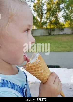 Une petite fille d'apparence caucasienne C tails assis sur un banc sur une journée ensoleillée et de manger un cornet de crème glacée Banque D'Images