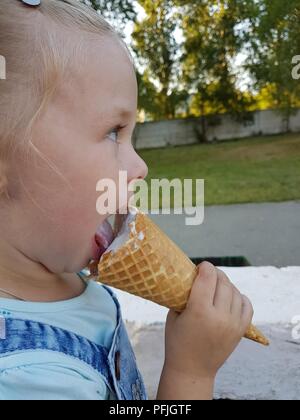 Une petite fille d'apparence caucasienne C tails assis sur un banc sur une journée ensoleillée et de manger un cornet de crème glacée Banque D'Images