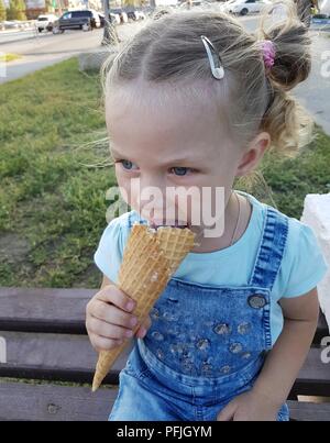 Une petite fille d'apparence caucasienne C tails assis sur un banc sur une journée ensoleillée et de manger un cornet de crème glacée Banque D'Images