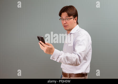 Jeune homme surpris en shirt looking at mobile phone while standing against gray background studio Banque D'Images