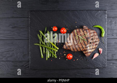 Fun pour de délicieux steaks de boeuf au barbecue avec des rayures sur la plaque en ardoise noire servi avec tomates, asperges grillées, d'ail et d'épices, vue d'en haut Banque D'Images