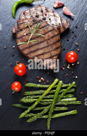 Fun pour de délicieux steaks de boeuf au barbecue avec bandes servi avec tomates, asperges grillées, d'ail et d'épices sur la plaque en ardoise noire, le sanglier en bois noir Banque D'Images