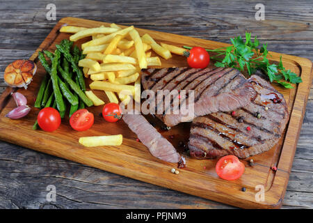 Fun pour de délicieuses grillades de bœuf juteux à moyen et des biftecks découpés en morceaux servis avec frites, tomates, asperges, tomates, ail et épices sur Cuttin ' Banque D'Images